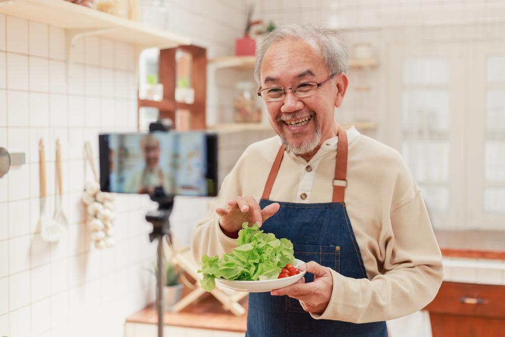 An older man with a salad dish is recording for a social media post. 